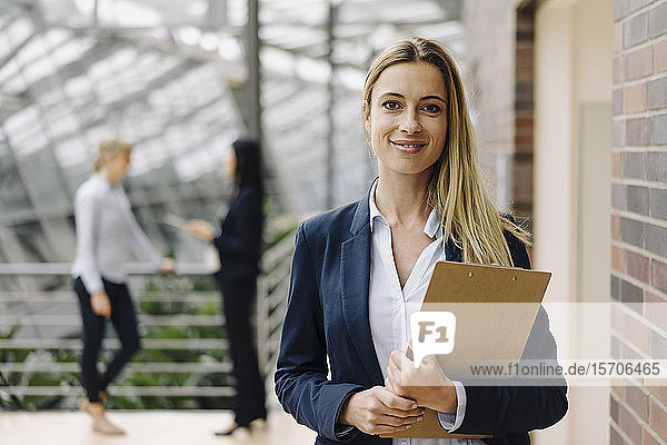 Porträt einer selbstbewussten jungen Geschäftsfrau in einem modernen Bürogebäude mit Kollegen im Hintergrund
