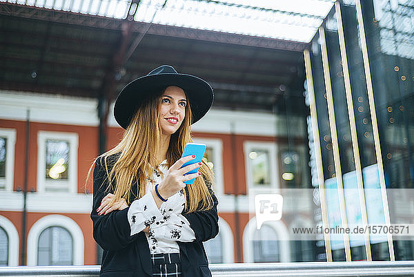 Lächelnde junge Frau mit Hut  die am Bahnhof mit dem Handy telefoniert