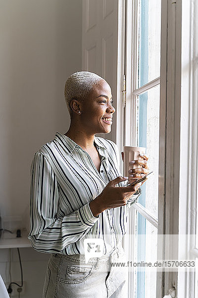 Happy woman with cell phone and coffee cup looking out of window