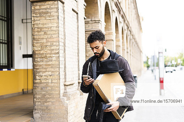 Junger Mann mit Hut und Paket schaut auf Handy in der Stadt