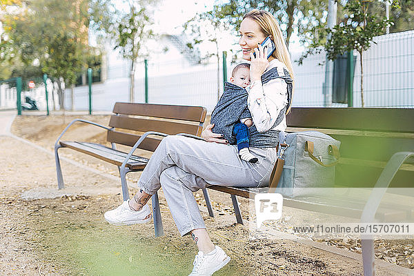 Glückliche Mutter ruht sich mit dem kleinen Jungen auf einer Parkbank aus und telefoniert