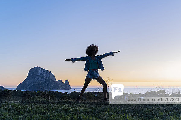 Junge Frau steht bei Sonnenuntergang auf einer Wiese und macht Yoga  Ibiza