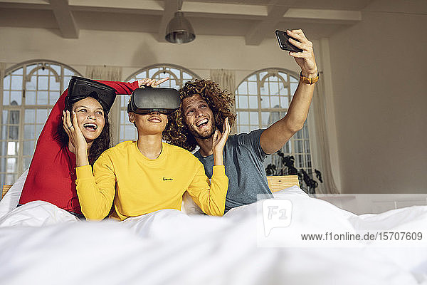 Glückliche Freunde die zu Hause im Bett mit VR-Brille und Smartphone Spaß haben