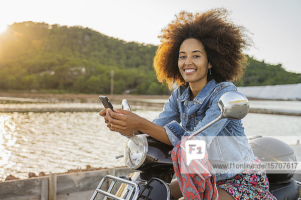 Young woman sitting riding on scooter and using smartphone at sunset  Ibiza