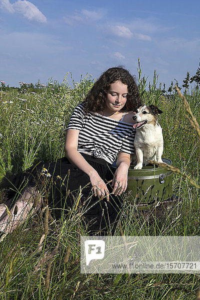 Porträt einer jungen Frau  die mit Koffern und ihrem kleinen Hund auf einer Wiese sitzt