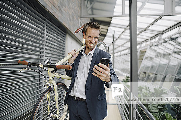 Lächelnder Geschäftsmann mit Handy mit Fahrrad in modernem Bürogebäude