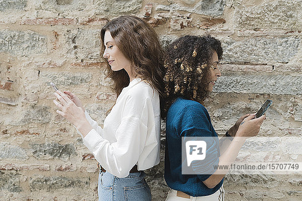 Two women standing back to back using smartphones
