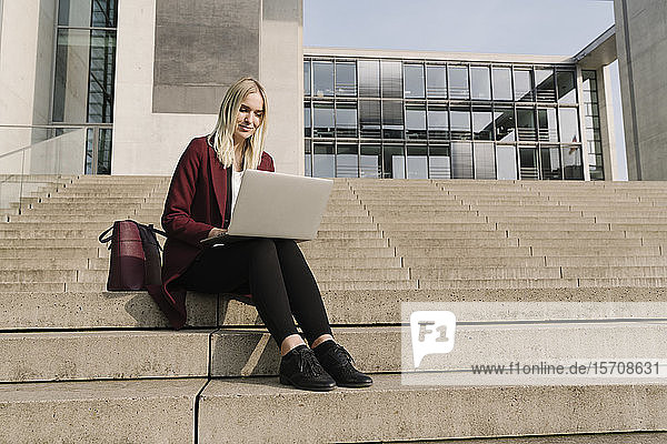 Blonde Geschäftsfrau mit Laptop im Hintergrund eines modernen Bürogebäudes