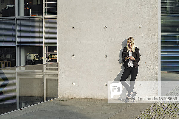 Blond businesswoman using smartphone leaning on wall