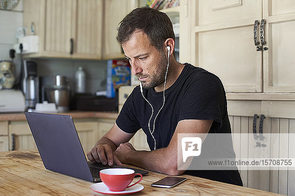 Man working from home  sitting at kitchen table  using laptop and smartphone