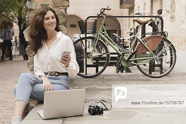 Smiling young woman with smartphone camera and laptop outdoors