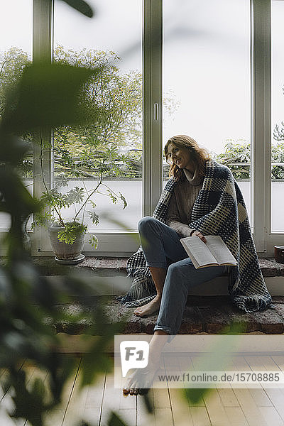 Eine reife Frau sitzt auf einer wunderbaren Fensterbank,  in eine Decke gewickelt,  liest ein Buch