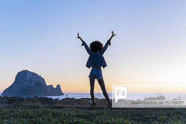Rückansicht einer jungen Frau  die bei Sonnenuntergang mit erhobenen Armen auf einem Aussichtspunkt steht  Ibiza