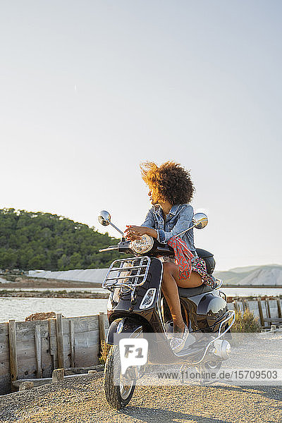 Junge Frau sitzt auf einem Motorroller und betrachtet den Sonnenuntergang  Ibiza