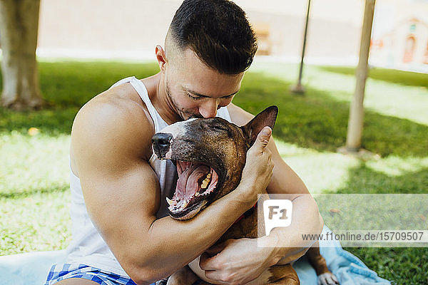 Muskulöser Mann sitzt auf einer Decke auf einer Wiese und umarmt seinen Hund