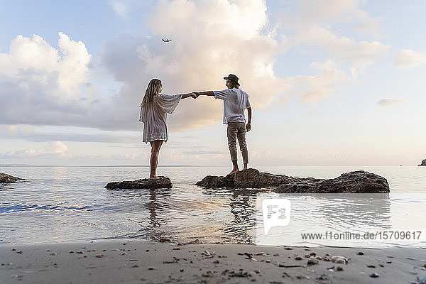 Junges Paar steht auf Felsen vor dem Meer und hält sich an den Händen  Ibiza  Balearen  Spanien
