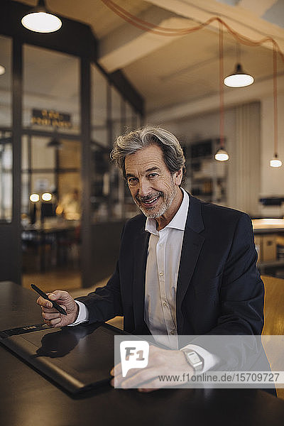Portrait of smiling senior businessman using graphics tablet at desk in office