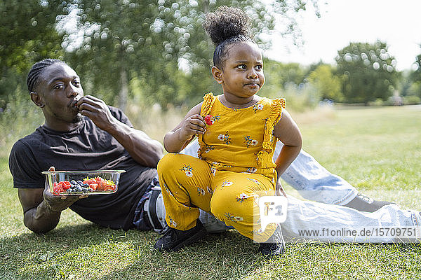 Vater und Tochter essen Obst auf einer Wiese in einem Park
