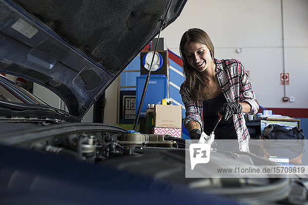 Junge Gelegenheitsfrau in Hemd und Handschuhen  die mit dem Auto im Reparaturdienst arbeitet und den Motorölstand kontrolliert