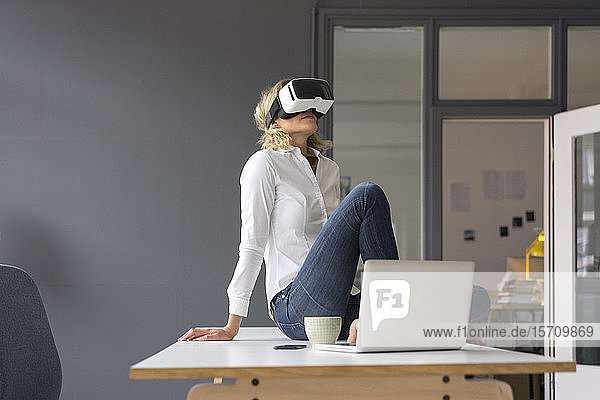 Junge Geschäftsfrau sitzt im Büro auf dem Schreibtisch und trägt eine VR-Brille
