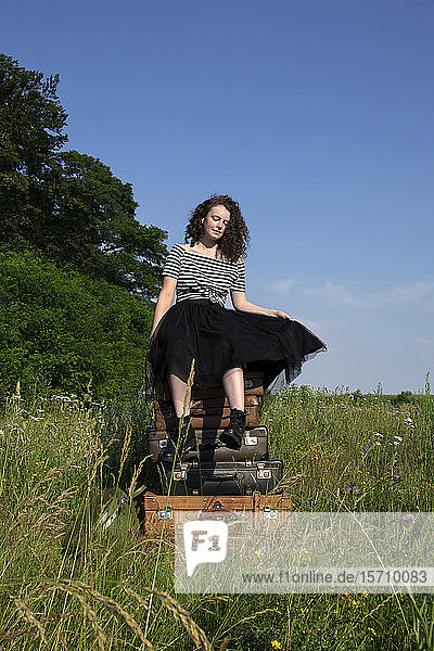 Porträt einer jungen Frau  die auf einem Stapel alter Koffer auf einer Wiese sitzt
