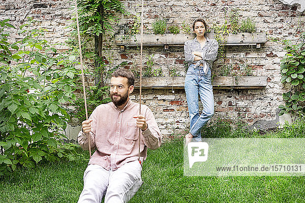 Couple in backyard  man sitting on swing  woman leaning on wall  looking displeased