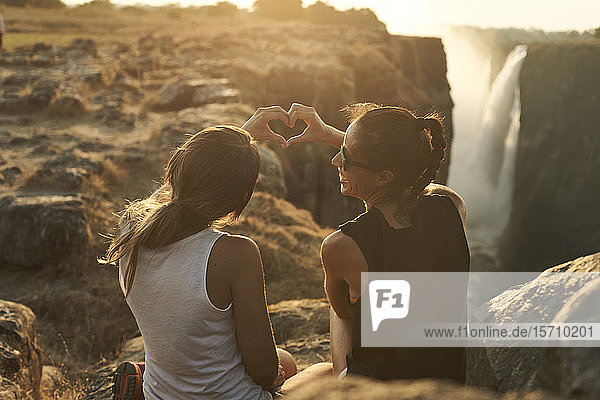 Frauen  die bei Sonnenuntergang vor den Viktoriafällen sitzen und ein Herz mit ihren Händen formen  Simbabwe