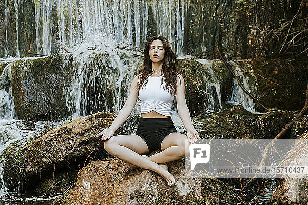 Junge Frau  die auf einem Wasserfall Yoga praktiziert  auf einem Felsen sitzend