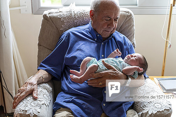 Großvater sitzt in einem Sessel und hält ein Neugeborenes