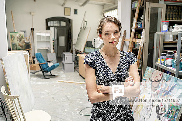 Junge Künstlerin steht mit verschränkten Armen in ihrem Atelier