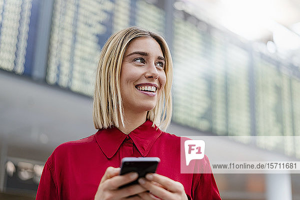 Lächelnde junge Geschäftsfrau mit Handy bei der Ankunft Abflugtafel am Flughafen