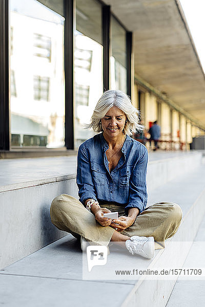Mature businesswoman sitting cross-legged on steps in the city  using smartphone