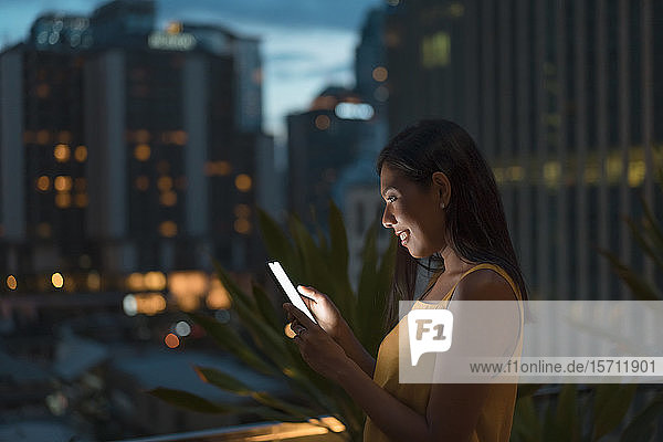Lächelnde Frau steht in der Abenddämmerung auf der Dachterrasse und schaut auf Handy  Bangkok  Thailand