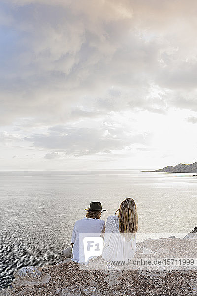 Back view of young couple sitting on rock in front of the sea  Ibiza  Balearic Islands  Spain