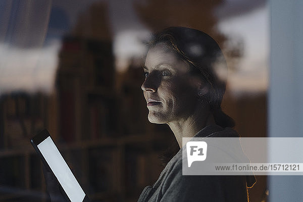 Frau  die in der Dämmerung aus dem Fenster schaut und ein digitales Tablet hält