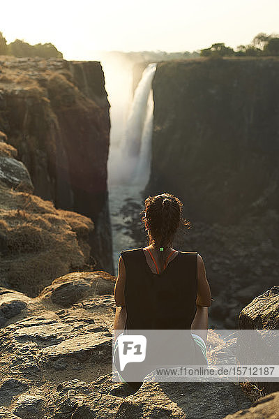 Frau sitzt auf der Spitze eines Felsens und genießt die Victoriafälle bei Sonnenuntergang  Simbabwe