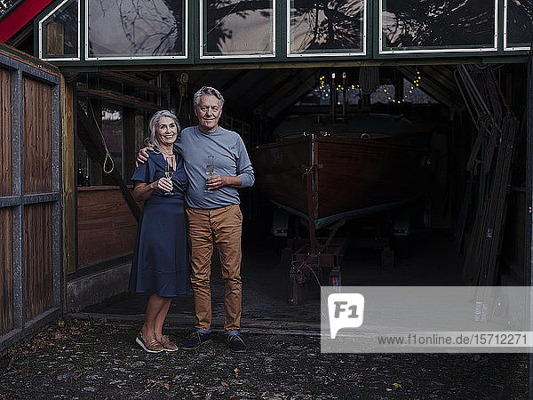 Senior couple standing in front of boathouse with glass of champagne