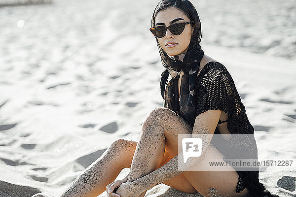 Junge Frau mit Kopftuch und Sonnenbrille am Strand sitzend