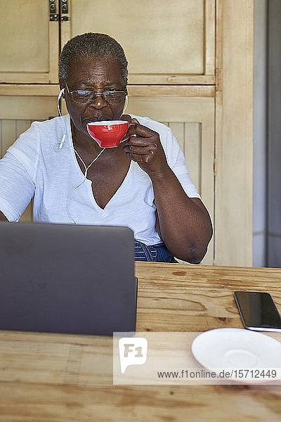 Ältere Frau sitzt am Küchentisch mit Laptop und trinkt aus der Tasse