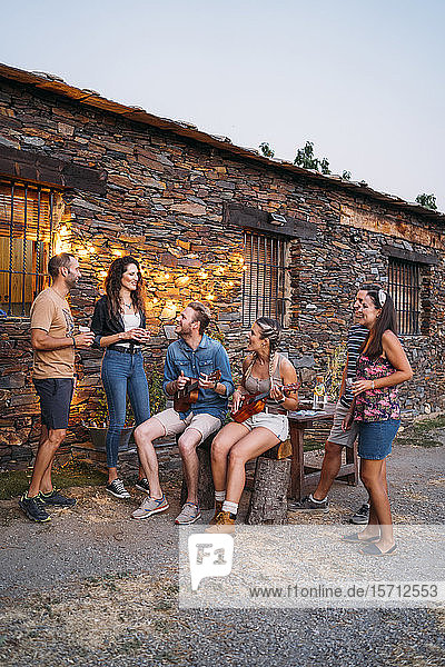 Gruppe von Freunden spielt in der Abenddämmerung Ukulele im Freien an einem Steinhaus