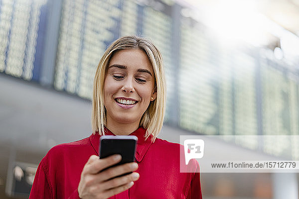 Lächelnde junge Geschäftsfrau benutzt Handy bei der Ankunft Abflugtafel am Flughafen
