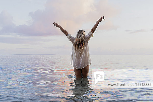 Rückansicht einer jungen Frau  die bei Sonnenuntergang eine weiße Bluse im Meer trägt