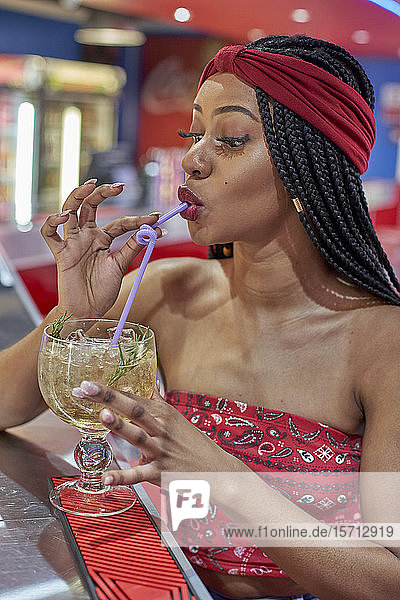 Junge Frau mit geflochtener Frisur sitzt an der Bar eines Restaurants und trinkt einen Cocktail mit einem Strohhalm