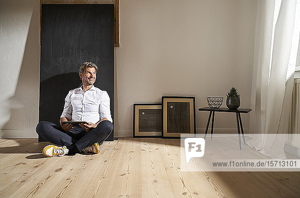 Entspannter  reifer Mann mit digitalem Tablett  der zu Hause auf dem Boden sitzt und aus der Ferne schaut