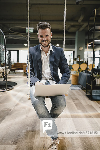 Geschäftsmann mit Laptop im Coworking Space  auf der Schaukel sitzend