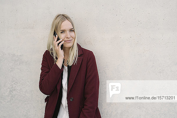 Blonde Geschäftsfrau mit Smartphone  an einer Wand lehnend  in die Kamera blickend