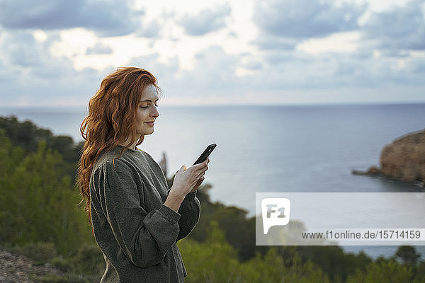Rothaarige junge Frau benutzt Handy an der Küste  Ibiza  Spanien