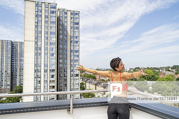 Rückenansicht einer glücklichen Frau  die mit ausgestreckten Armen auf der Dachterrasse steht