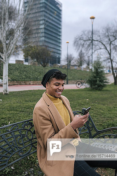 Lächelnder junger Mann sitzt auf einer Bank und benutzt sein Handy