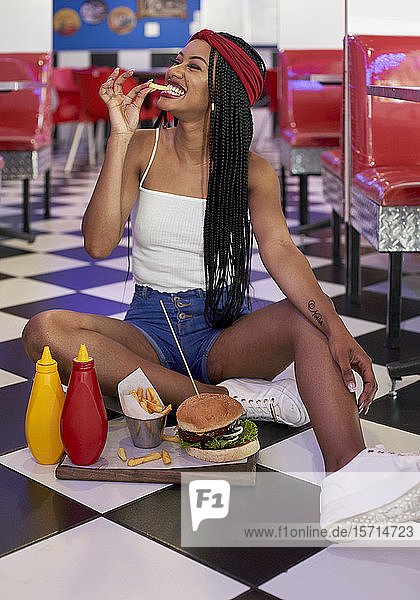 Junge Frau mit geflochtener Frisur sitzt mit ihrem Hamburgerteller auf dem Boden und isst einen Chip